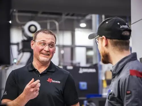 Technician in safety glasses speaks with a coworker beside CNC equipment on a manufacturing floor.